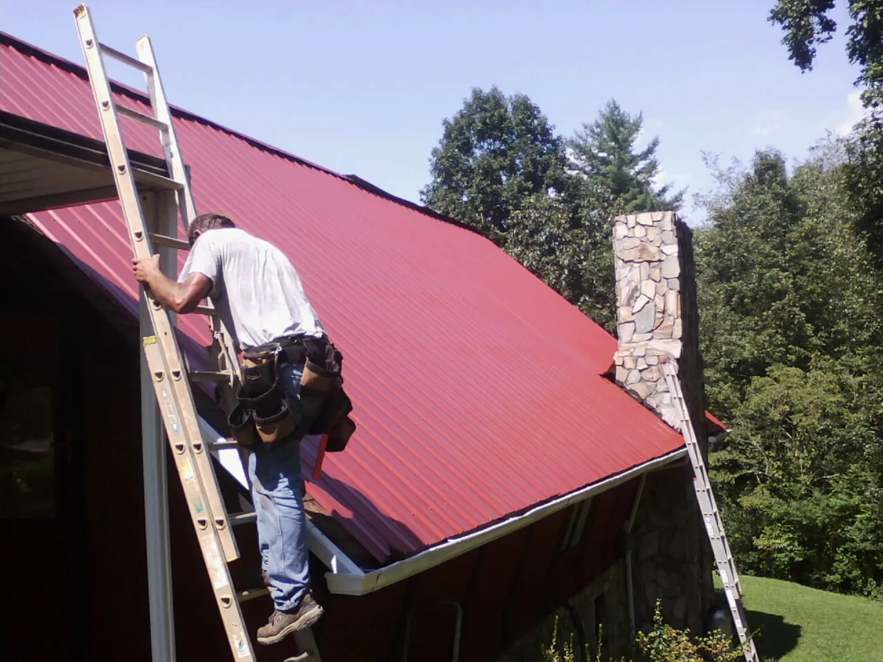 Roofer on a ladder beside a red metal roof and stone chimney — Soffit & Fascia Repair by Roofing Brothers in Killbuck