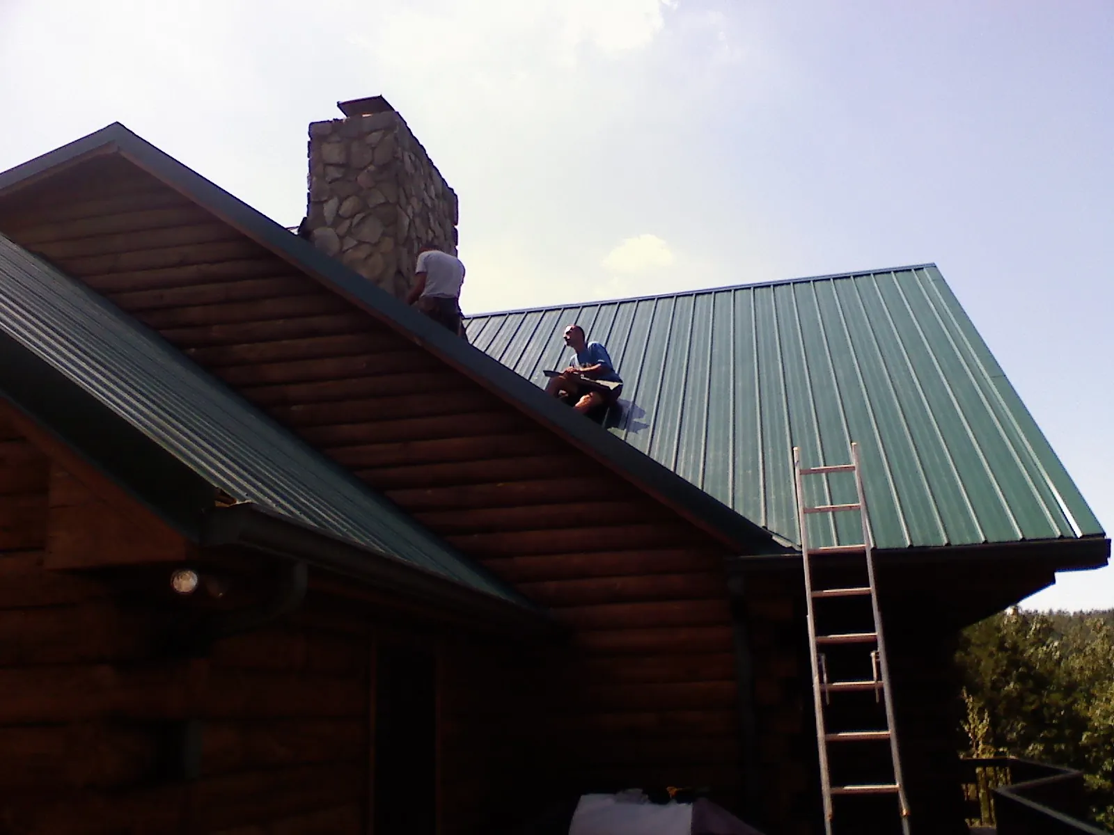 Roofers installing green metal panels beside a stone chimney on a log home in Killbuck